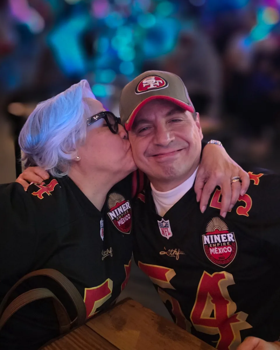 Miembro recibiendo un beso en la mejilla, ambos con jerseys negros de Niner Empire México